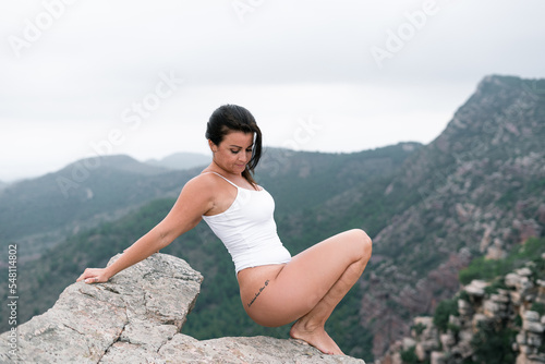 Caucasian mature woman in a white swimsuit calm and relaxed exercising on top of a big rock