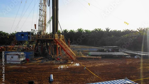 Cinematic 4K Drone Footage of Onshore Drilling Rig equipment structure for oil exploration and exploitation in the middle of jungle surrounded by palm oil trees during sunset and high oil price. photo