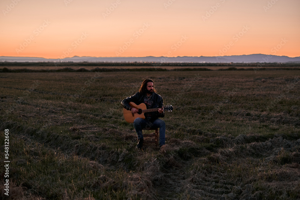Caucasian young man with a beard and long hair sitting on a small stool calmly playing the guitar at sunset in a lonely place