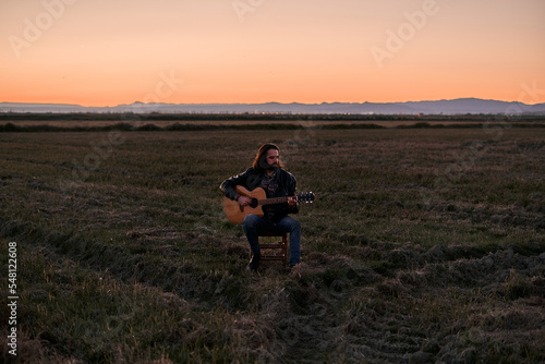 Caucasian young man with a beard and long hair sitting on a small stool calmly playing the guitar at sunset in a lonely place