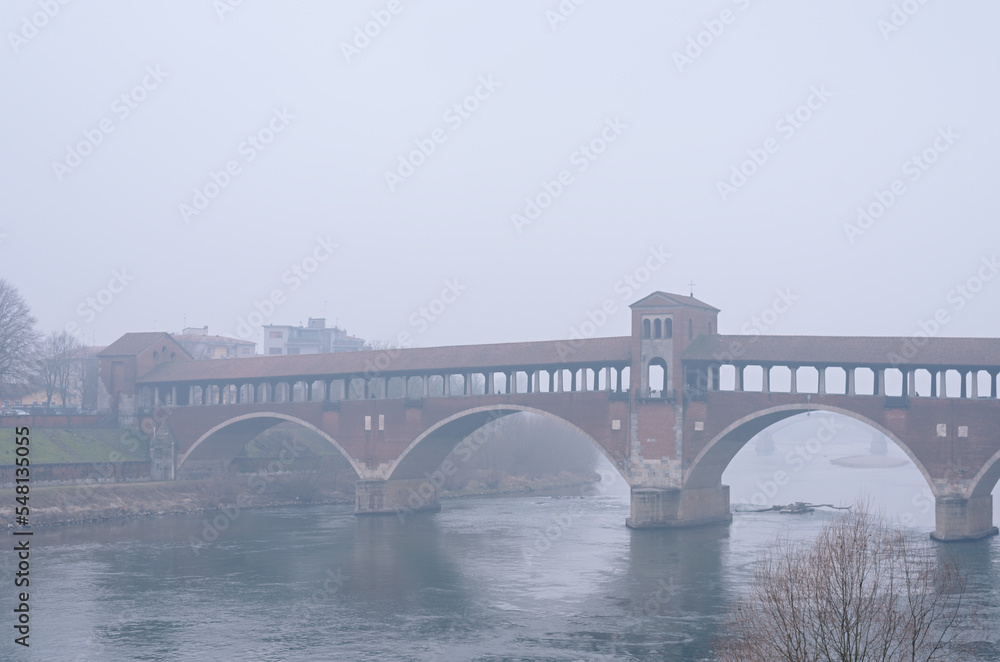 Fototapeta premium Ancient covered bridge surrounded by fog in winter. Pavia. Italy