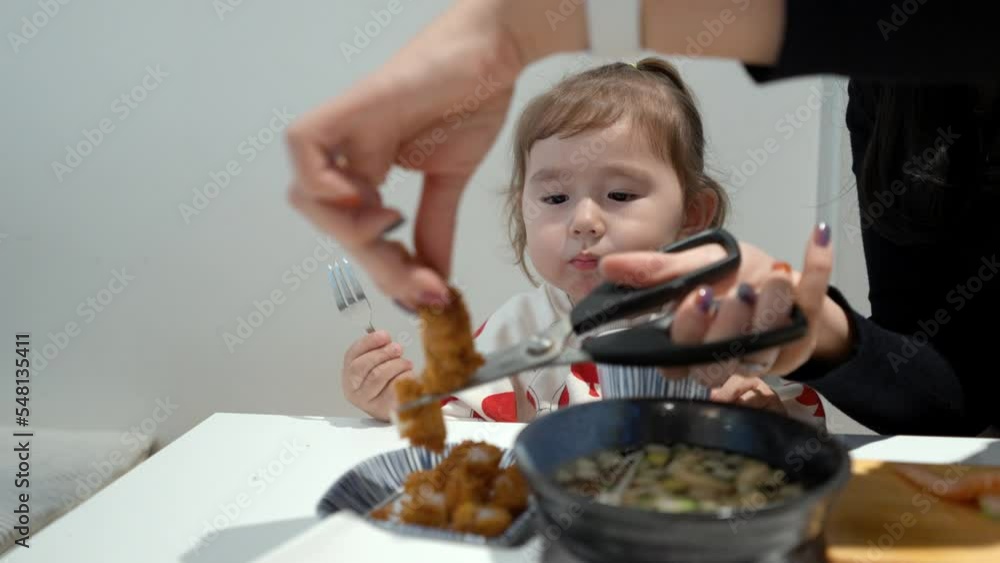 Mother's Hands Cutting Fried Fish Fillet Into Bite Size Pieces. Beautiful Female Toddler Munching Food While Watching. medium shot Mother's Hands Cutting Fried Fish Fillet Into Bite Size Pieces. Beautiful Female Toddler Munching Food While Watching. medium shot