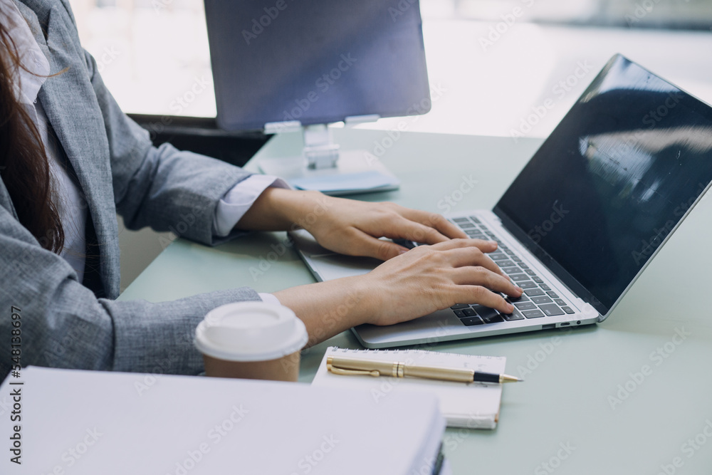 Fototapeta premium Young business woman sitting in office at table and using smartphone. On desk is laptop and tablet computer, on screen charts and graphs. Woman analyzing data. Student learning online.