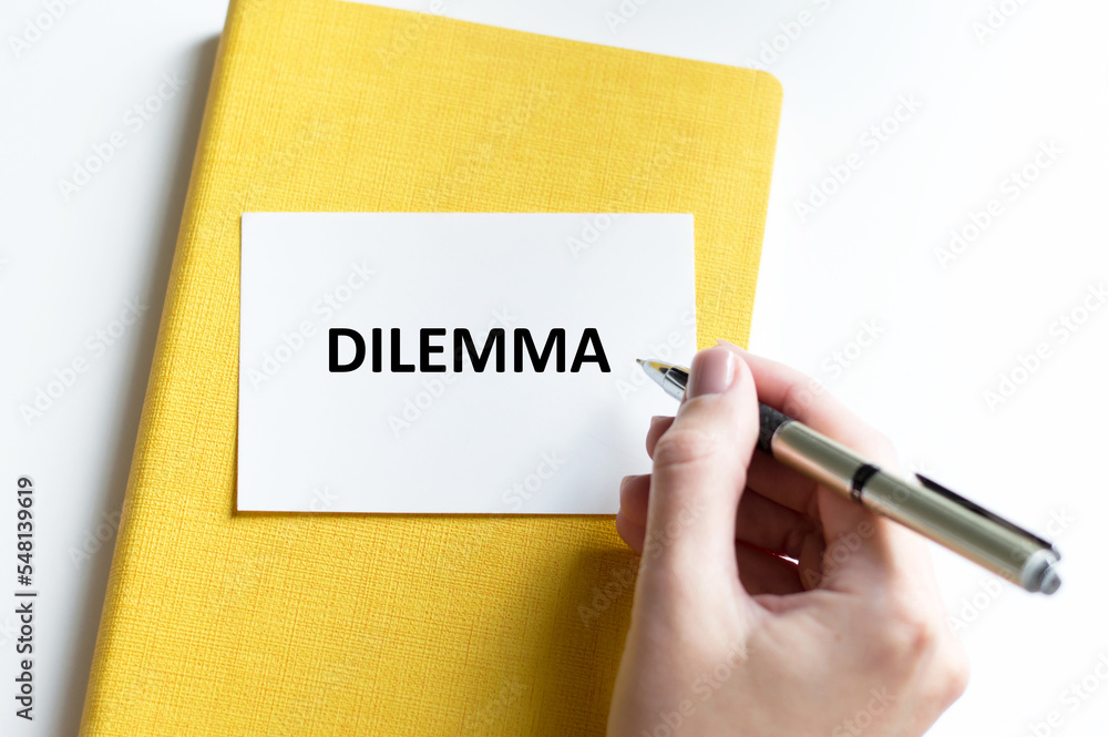 Text DILEMMA on the card next to the hand of a person on the table, writes the text Text DILEMMA on the card next to the hand of a person on the table, writes the text