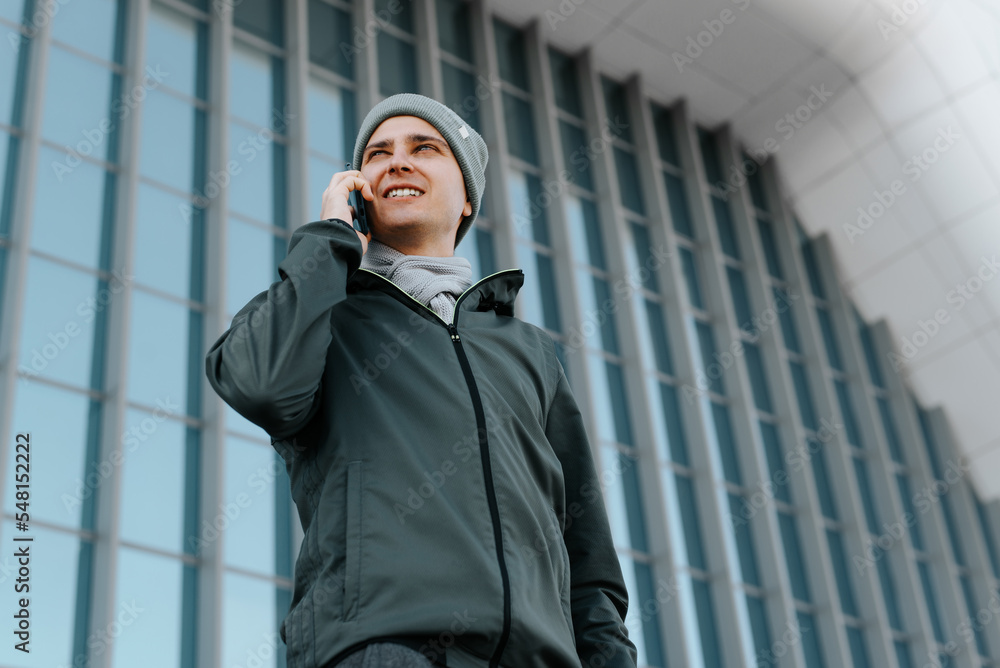 Smiling young man talking on a mobile phone while standing against backdrop of modern geometric architecture and looking away. Portrait of guy in outerwear calling on smartphone outdoors Smiling young man talking on a mobile phone while standing against backdrop of modern geometric architecture and looking away. Portrait of guy in outerwear calling on smartphone outdoors