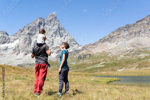 Young couple with toddler standing close to a mountain lake while pointing and looking at majestic Matterhorn since Cervinia in Italian Alps.