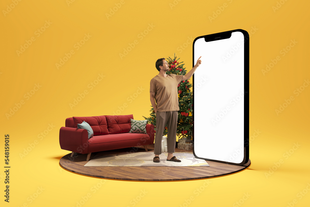 Christmas atmosphere at home. Photo and 3d illustration of man standing next to huge 3d model of smartphone with empty white screen isolated on yellow background. Mockup for ad - obrazy, fototapety, plakaty 