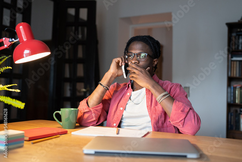 Stressed puzzled African American man making phone call as found out about overdue work deadlines. Shocked depressed guy freelancer in casual clothes sits at table with laptop and mug in apartment