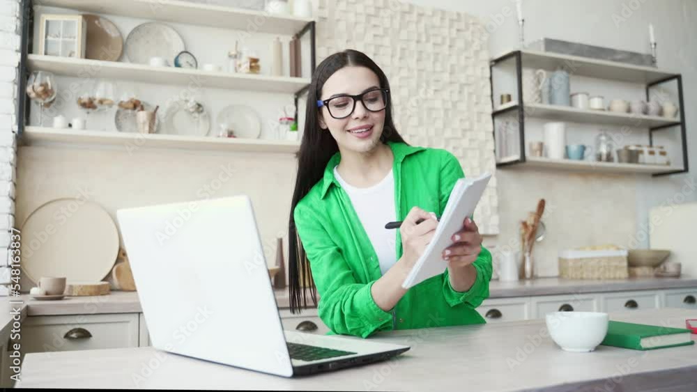 Portrait of dark haired student with eyeglasses have a remote lesson, talking with teacher, showing notebook and looking at the laptop camera in the domestic kitchen. Distance education concept Portrait of dark haired student with eyeglasses have a remote lesson, talking with teacher, showing notebook and looking at the laptop camera in the domestic kitchen. Distance education concept