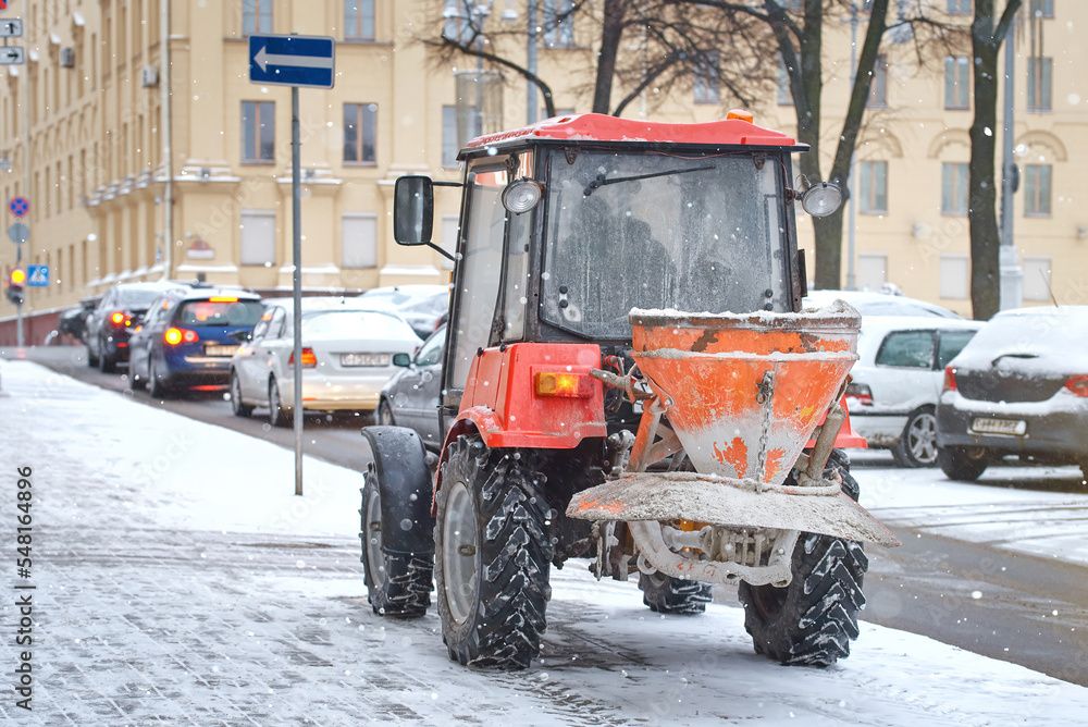 Tractor with salt spreader in winter day during blizzard, tractor ready sprinklr rock salt and sand on sidewalk, winter road maintenance, prevent slipping.Tractor de-icing street, melt ice and snow Tractor with salt spreader in winter day during blizzard, tractor ready sprinklr rock salt and sand on sidewalk, winter road maintenance, prevent slipping.Tractor de-icing street, melt ice and snow