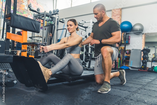 Young, athletic, caucasian woman performing seated back pull on the machine and her man coach helping her to do the exercise correctly in the gym. High quality photo