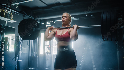 A Professional Female Powerlifter Training with Strength Exercices Using a Barbell in a Dark Empty Gym. A Hardworking Sports Woman Works Out, Lifts Weights with Endurance