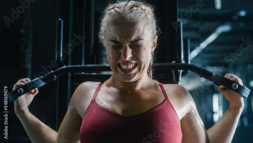 An Agile Future Champion Training by Using Lat Pull Down Machine and Sweating in a Dark Gym. The Portrait of a Focused Sports Woman Doing Workout Exercises with an Expressive Face