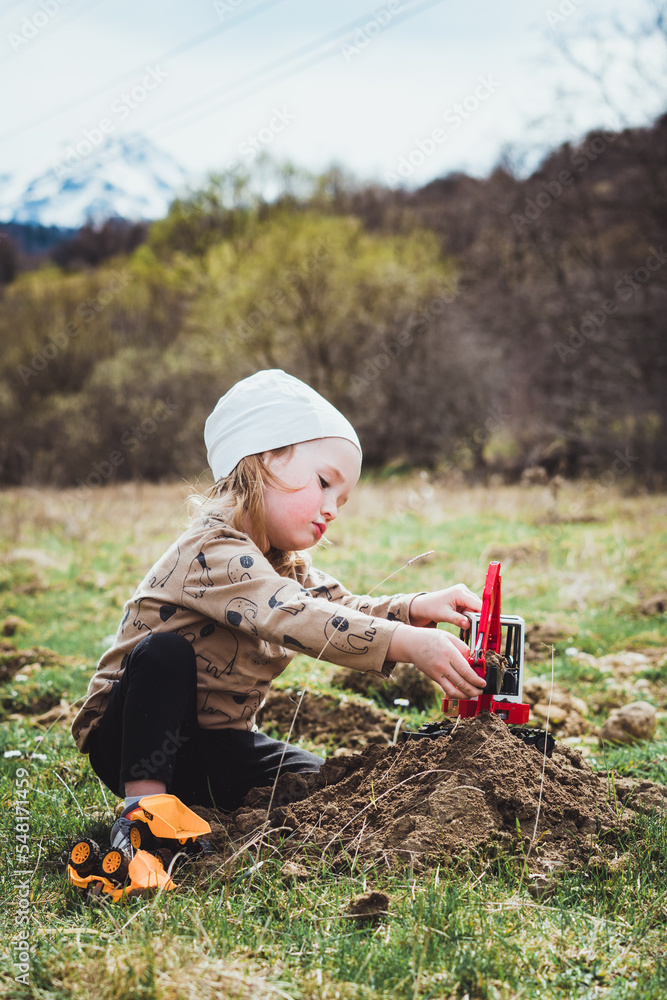 Beautiful young boy child with long blond hair playing construction site in the dirt with diggers and dumpers while out in nature. Free and independent play while hiking in a natural park.