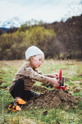 Beautiful young boy child with long blond hair playing construction site in the dirt with diggers and dumpers while out in nature. Free and independent play while hiking in a natural park.