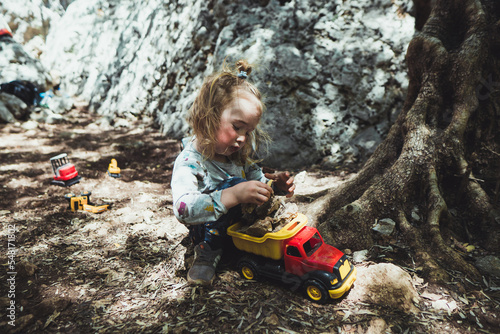 Beautiful young boy child with long blond hair playing construction site in the dirt with diggers and dumpers while out in nature. Free and independent play while hiking in a natural park.