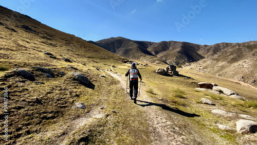 A man with trekking sticks and a backpack walks along a hiking trail among the mountain peaks. A lonely tourist travels high in the mountains on a sunny summer day. Independent hike in the mountains.