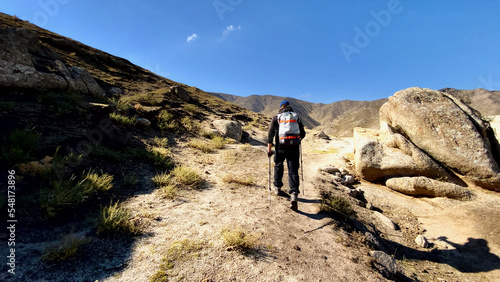 A man with trekking sticks and a backpack walks along a hiking trail among the mountain peaks. A lonely tourist travels high in the mountains on a sunny summer day. Independent hike in the mountains.
