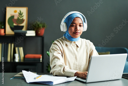 Young Muslim female student with headphones sitting in front of laptop by workplace while listening to teacher during online lesson photo