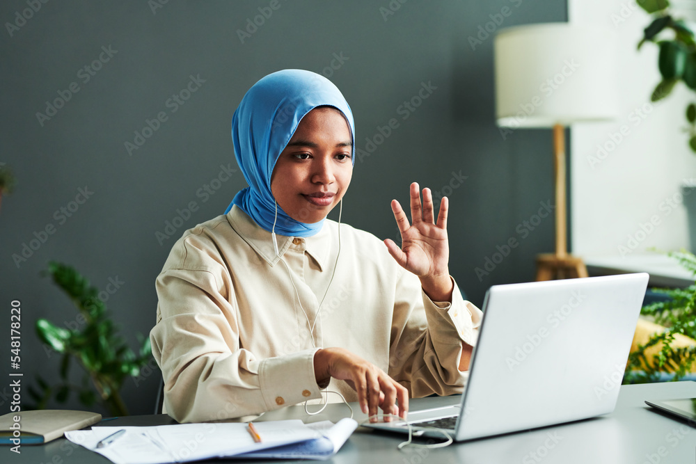 Young Muslim student in blue hijab greeting teacher by waving hand during online lesson while sitting by desk in front of laptop Young Muslim student in blue hijab greeting teacher by waving hand during online lesson while sitting by desk in front of laptop