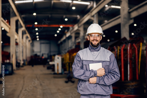 A factory supervisor in work wear is holding tablet and smiling at the camera in facility. © dusanpetkovic1