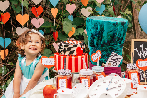 Little smiling girl 2 years old dressed as Alice in Wonderland near table with fairy tale props. Cake, Hatters hat, inscriptions Eat me and Drink me, clock, card suits. Themed childrens book birthday