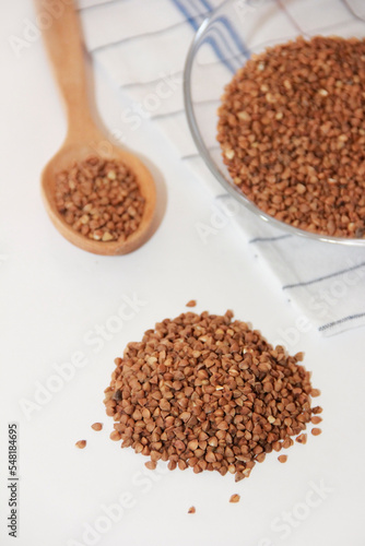small grains of brown natural dry buckwheat in a transparent glass plate and in a wooden spoon