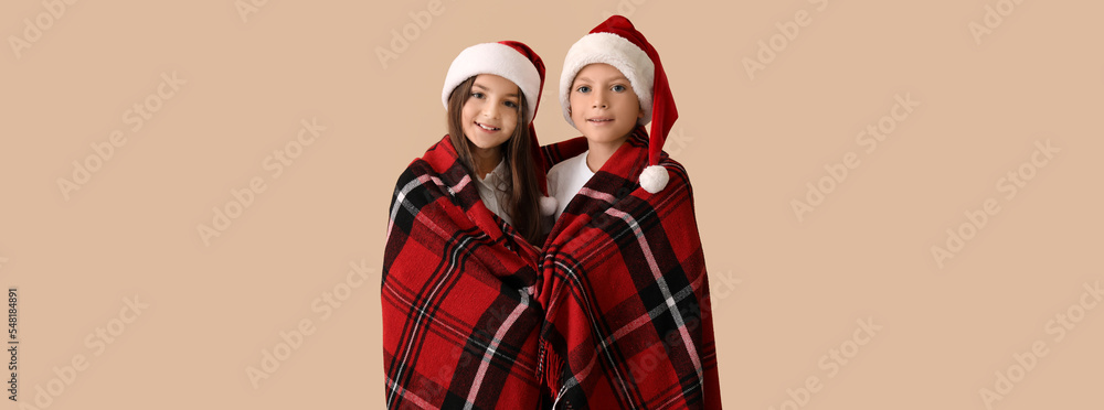 Cute children in pajamas and Santa hats wrapped in plaid on beige background
