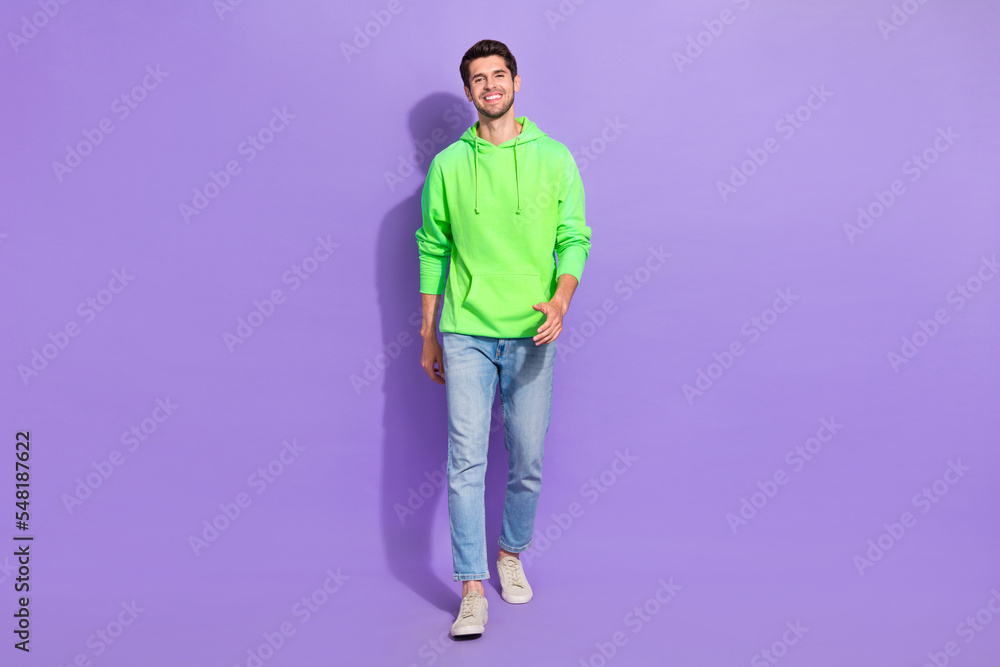 Full size photo of handsome cheerful positive guy dressed green hoodie denim jeans walking street isolated on violet color background Full size photo of handsome cheerful positive guy dressed green hoodie denim jeans walking street isolated on violet color background