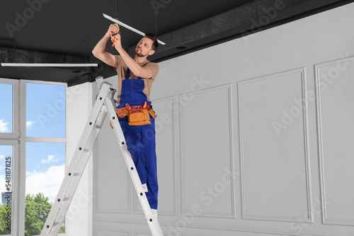 Electrician in uniform installing ceiling lamp indoors. Space for text