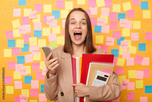 Photo of surprised happy pleased young adult woman holding paper documents and mobile phone, looking at camera screaming happily, posing against yellow wall with colorful sticky post notes.