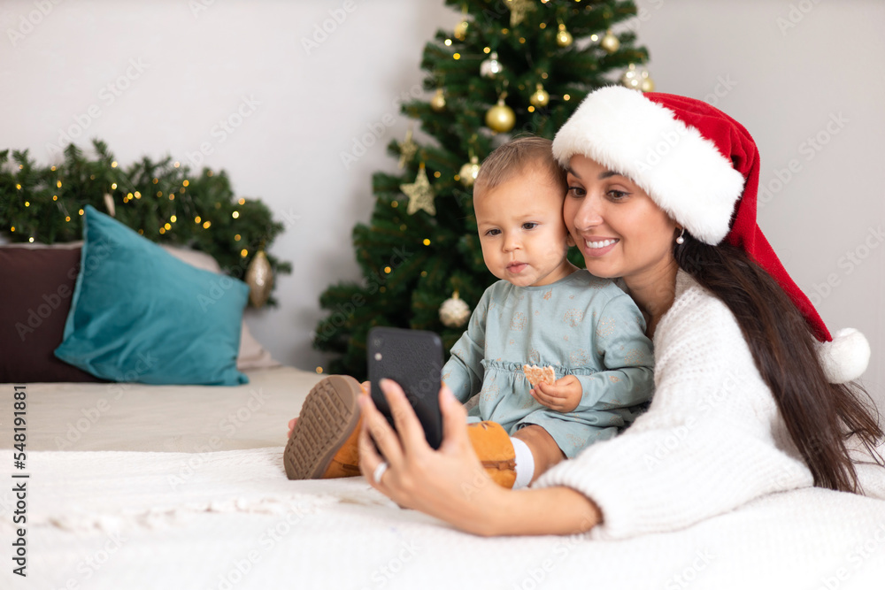 Merry Christmas. A beautiful mother and a little daughter call their relatives via video link or take a selfie on a smartphone. Cozy decorated bedroom. Lifestyle Merry Christmas. A beautiful mother and a little daughter call their relatives via video link or take a selfie on a smartphone. Cozy decorated bedroom. Lifestyle