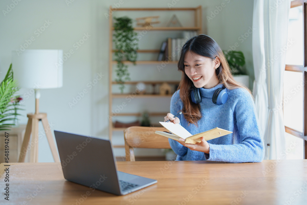 Portrait of a teenage Asian woman using a computer, wearing headphones and using a notebook to study online via video conferencing on a wooden desk in library Portrait of a teenage Asian woman using a computer, wearing headphones and using a notebook to study online via video conferencing on a wooden desk in library