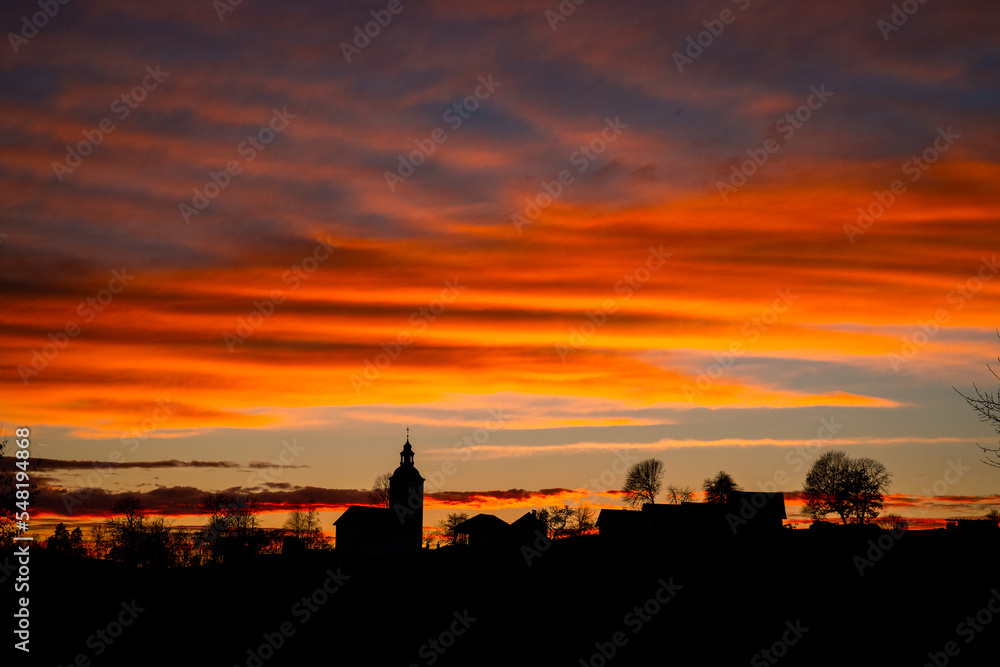 Silhouette of a village in slovenia, black silhouette of a church, houses and trees in front of a beautiful red sunset and majestic cloudy skies.