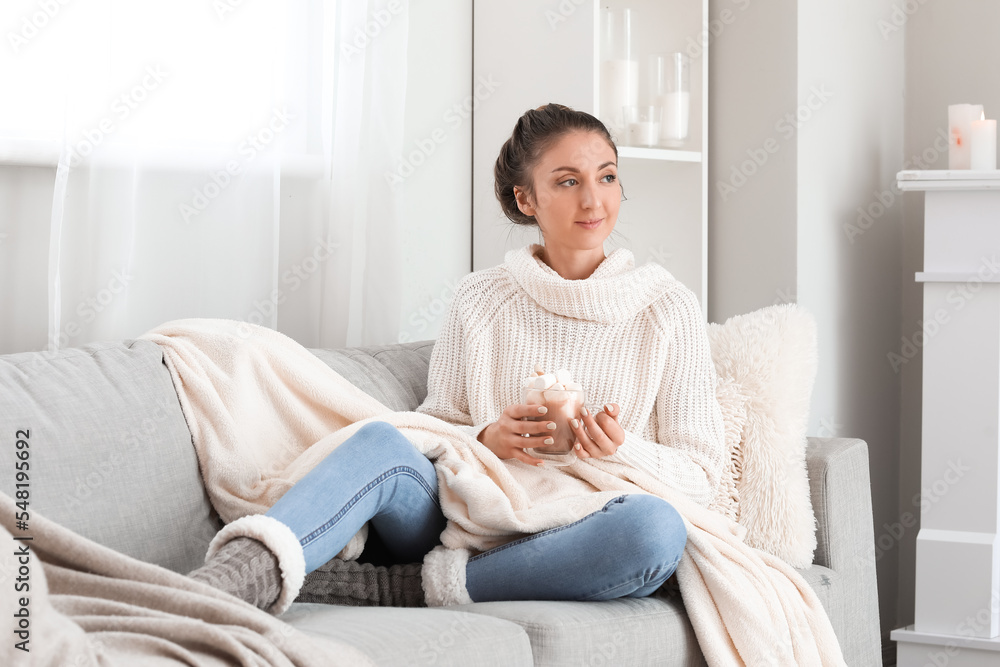 Young woman with glass cup of cocoa and plaid sitting on sofa at home