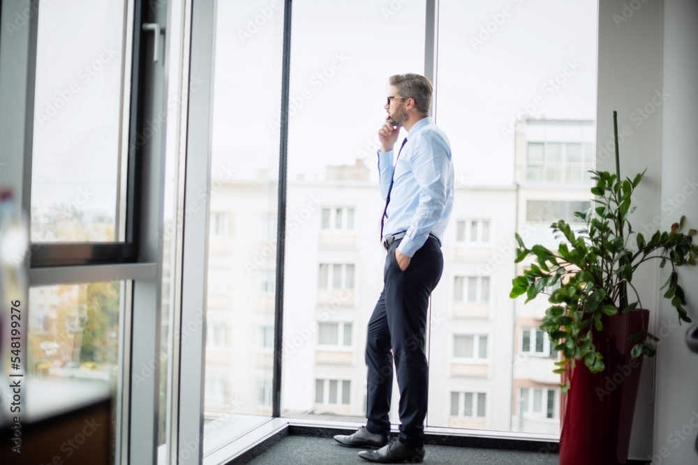 Full length of thinking businessman looking out of office over city. Confident man wearing glasses and business casual. Full length of thinking businessman looking out of office over city. Confident man wearing glasses and business casual.