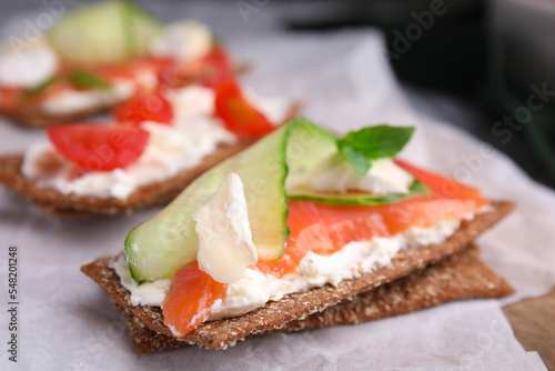 Tasty rye crispbreads with salmon, cream cheese and cucumber on parchment paper, closeup