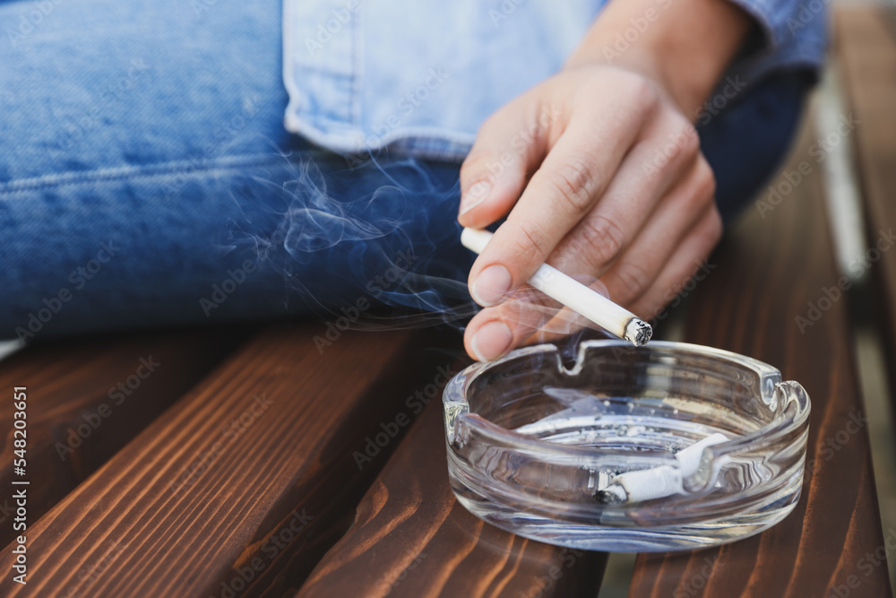 Woman holding cigarette over glass ashtray on bench outdoors, closeup