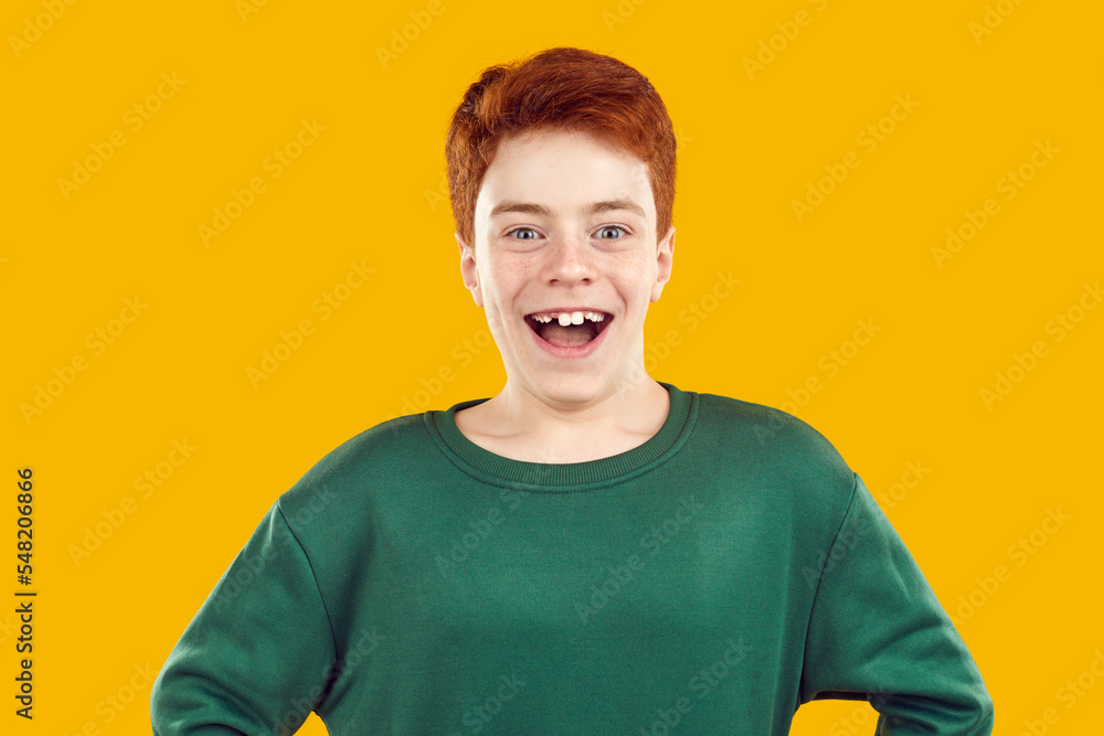 Portrait of cheerful preteen boy who laughs funny showing smile with crooked teeth. Caucasian red-haired boy smiling joyfully, isolated on orange background. Concept of teeth alignment in childhood.