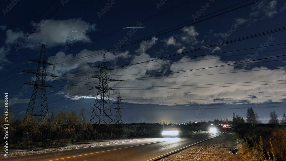 Night suburban highway under power lines. Dark blue sky with white clouds, asphalt road without lighting. Cars are driving along the road with their headlights on, leaving a long trail of light.
