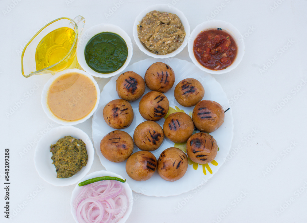 Litti in white plate with tomato, brinjal, potato, pointed gaurd and coriander chutney or chokha and ghee. It is popular in Bihar, Uttar Pradesh and Jharkhand. White background. top view
