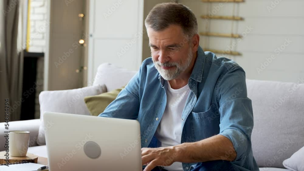 Senior old mature man typing on laptop browsing internet using apps sit on sofa alone Senior old mature man typing on laptop browsing internet using apps sit on sofa alone