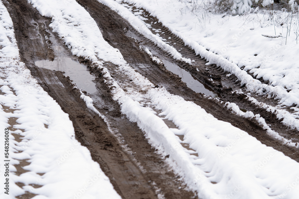 Old broken road with pits and puddles through winter forest.