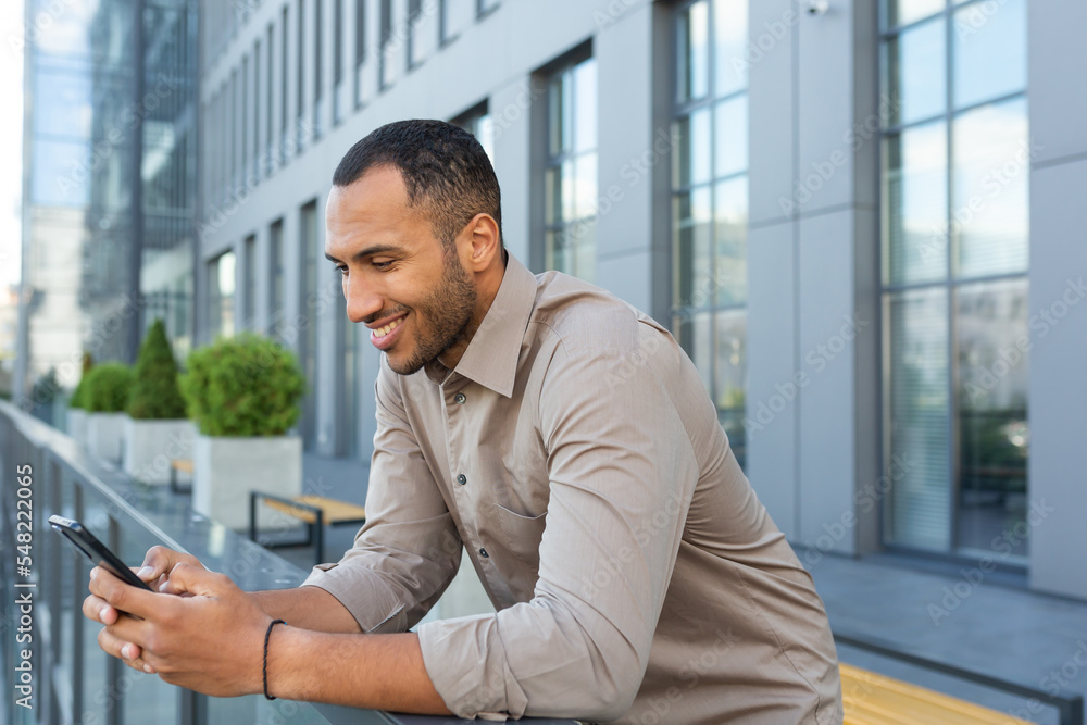 A young handsome Latin American man is standing on the veranda of the office center, holding a phone in his hands, typing a message, writing a text, using it. A young handsome Latin American man is standing on the veranda of the office center, holding a phone in his hands, typing a message, writing a text, using it.