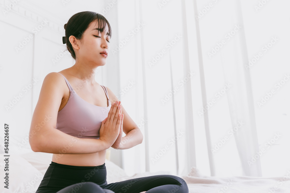 Young attractive sporty Asian woman practicing yoga on the bed, doing Ardha Padmasana exercise, meditating in Half Lotus pose with namaste, indoor working out at home, wearing sportswear.