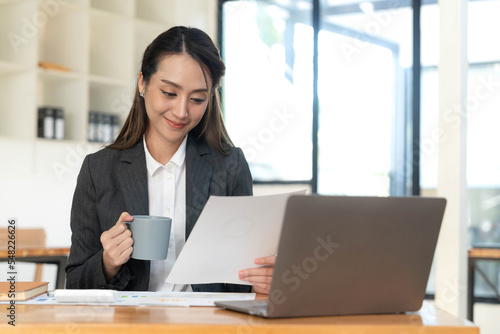 Beautiful young Asian businesswoman smiling holding a coffee mug and laptop working at the office.
