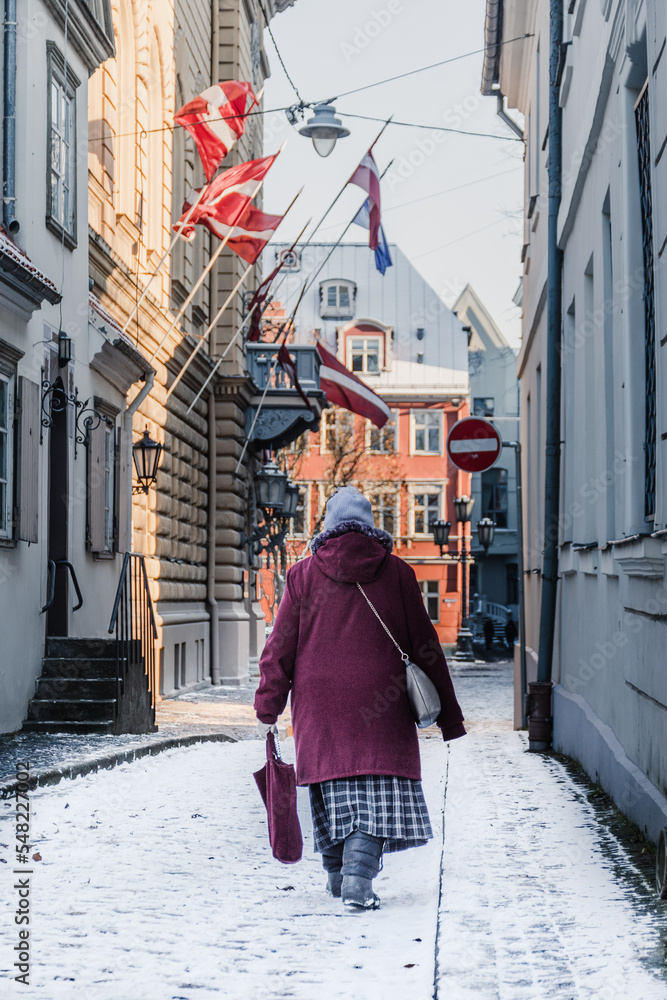Elderly woman wearing winter warm clothes walking in a narrow street covered by the snow in Riga Old Town near Parliament House