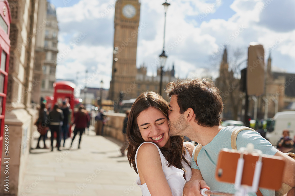 Couple, travel and selfie with kiss outdoor in London for romantic holiday, adventure and love in city street. Man, woman and smartphone, happiness with smile in traveling photo, tourist and romance.