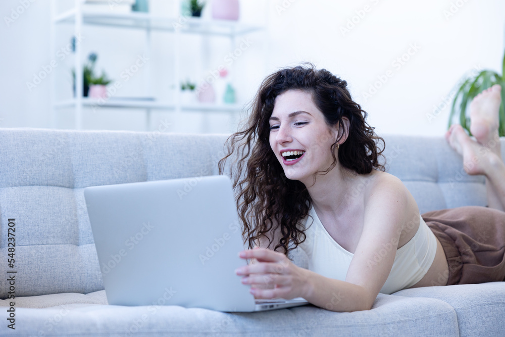 young student lies on sofa with a big smile while browsing the internet on her computer young student lies on sofa with a big smile while browsing the internet on her computer