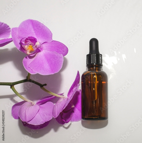 Dark brown bottle of skin serum with dropper on gray background with purple orchid flowers, close-up